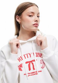 Biała bluza z kapturem z czerwonym tekstem graficznym i ilustracjami. Materiał wydaje się miękki, ma luźny krój i regulowany kaptur.