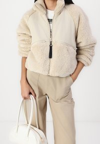 Beżowa kurtka na zamek z puszystą, teksturowaną dolną częścią i gładką górną częścią. Posiada czarną zawieszkę przy zamku. W zestawie z beżowymi spodniami i białą torbą.