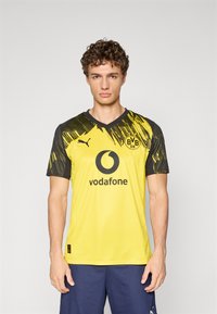 Geltonos ir juodos spalvos trumpomis rankovėmis futbolo marškinėliai su V formos iškirpte, kuriuose yra logotipas, rėmėjo tekstas ir abstraktus grafikos dizainas ant pečių.