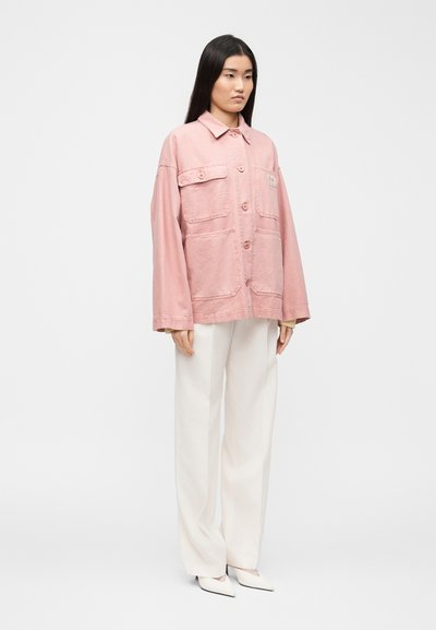 Γυναίκα με μακριά σκουρόχρωμα μαλλιά φοράει ροζ φαρδιά σακάκι με κουμπιά, λευκό παντελόνι φαρδύ στα πόδια και λευκά μυτερά παπούτσια, στέκεται μπροστά σε απλό φόντο.