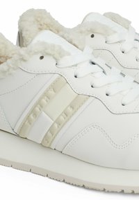 White sneakers with a textured faux fur collar, smooth leather upper, and embossed "TOMMY JEANS" logo on a cream stripe. Durable rubber sole.