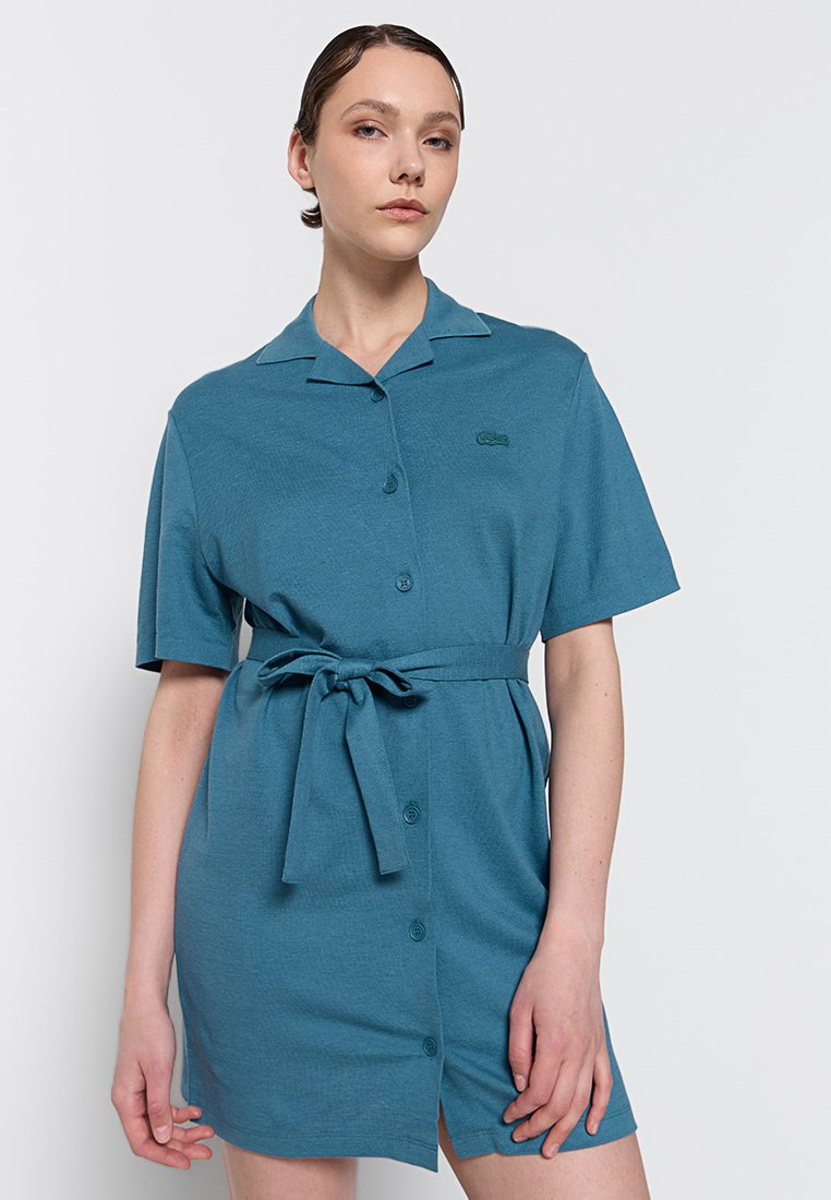 Niebieska sukienka z krótkim rękawem, z guzikiem z przodu, z paskiem do związania w talii, z małym zielonym haftowanym logo, wykonana z teksturowanej tkaniny.