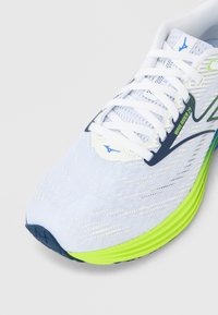 Běžecká bota s bílým síťovaným svrškem, zelenými a modrými akcenty, zaoblenou špičkou a texturovanou podrážkou pro lepší přilnavost. Má bílé tkaničky a značkové detaily.