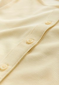 Tessuto giallo chiaro a coste con una fila di quattro bottoni di plastica sul davanti. Tessitura liscia che forma pieghe morbide.