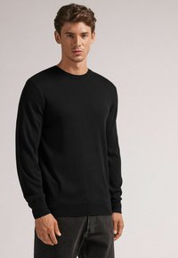 Svart crew-neck tröja i mjukt tyg. Ribbstickade manschetter och nederkant. Modellen bär tröjan med mörka jeans, vilket visar en slimmad design och slät textur.