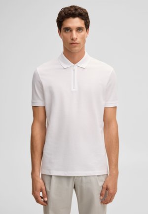 Młody mężczyzna z kręconymi włosami w białym, krótkim rękawie koszulce polo z zamkiem z przodu i jasnych, teksturowanych spodniach.