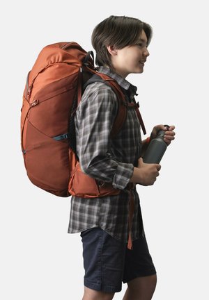 Orange backpack with adjustable straps, ergonomic design, and mesh back panel. User holds a gray water bottle, wearing a plaid shirt and shorts.