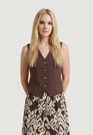 Donna bionda con capelli lunghi che indossa un gilet marrone con bottoni sopra una gonna marrone e bianca a fantasia, in piedi davanti a uno sfondo uniforme.