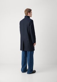 Cappotto sartoriale blu con colletto alto, caratterizzato da una silhouette dritta e uno spacco posteriore, indossato sopra jeans blu larghi e scarpe nere.