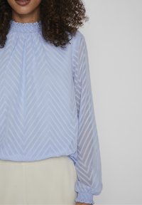 Světle modrá halenka s dlouhými rukávy, chevronovým vzorem, nařaseným výstřihem a elastickými manžetami. Volný střih, částečně průhledné rukávy a hladká tkanina.