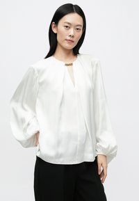 Blouse en soie blanche avec manches bouffantes, encolure en forme de goutte et un accent doré au col. Texture lisse qui complète la coupe ample.