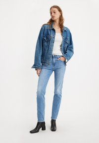 Modra jeans jakna z oversized krojem, svetlo modri jeans in črne gležnjarje na peto. Model nosi belo rebrasto majico brez rokavov.