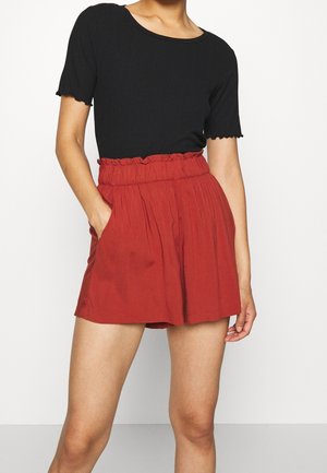 Vrouw die een zwart kortmouwtopje draagt en losse rode shorts met een hoge taille en zakken, staand tegen een effen achtergrond.