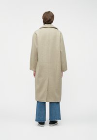 Μπεζ μάλλινο παλτό με φαρδύ γιακά, ίσια γραμμή, μακριά μανίκια και διάκοσμο στην πλάτη, φορεμένο πάνω από μπλε φαρδιά τζιν και μαύρα αθλητικά παπούτσια.