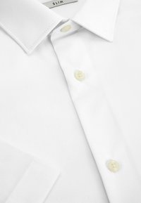 Bijela slim-fit košulja s oštrim kragnom. Ima biserno obojene gumbe i glatku pamučnu tkaninu. Bezručasto izrađeni ramena. Dupli manžet.