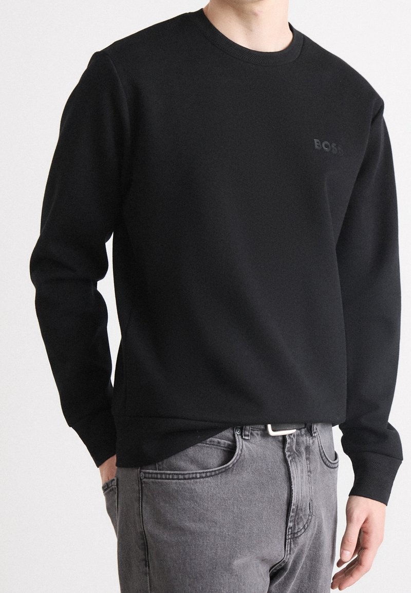 Felpa nera con collo rotondo, realizzata in cotone, con un logo goffrato sottile e polsini a costine, abbinata a jeans grigi.