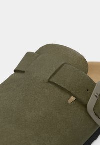 Olijfgroene suède klomp met een enkele verstelbare band en metalen gesp, met een gestructureerd oppervlak en een natuurlijke kurken voetbed.