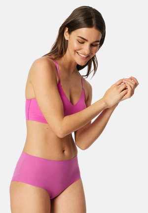 Vrouw die glimlacht en haar handen aanpast, gekleed in een felroze bralette en bijpassende hoge tailleondergoed, tegen een effen achtergrond.