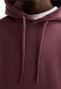 Primer piano di una persona che indossa un hoodie marrone bordeaux con cordini sopra una maglietta bianca a girocollo, mostrando la zona del collo e della parte superiore del petto.