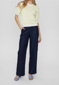 Žena oblečená v bílo-světle zeleném pruhovaném tričku s krátkým rukávem, tmavě modrých širokých džínech a světle modrých otevřených lodičkách, postávající s jednou rukou v kapse.