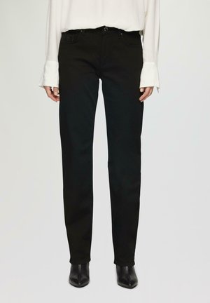 Jean droit noir en denim, avec un design classique à cinq poches et une texture subtile à la taille. Porté avec un blouse blanche.