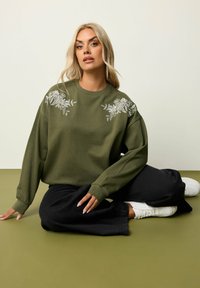 Felpa verde oliva con ricami floreali sulle spalle, abbinata a pantaloni neri e sneakers bianche. Tessuto morbido, vestibilità rilassata, maniche lunghe.