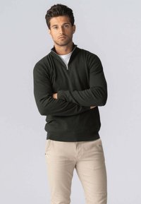 Mørkegrøn sweater med lynlås ved halsen, lavet af strikstof med ribbede manchetter og kant, kombineret med lysebeige bukser, sat mod en ensfarvet baggrund.