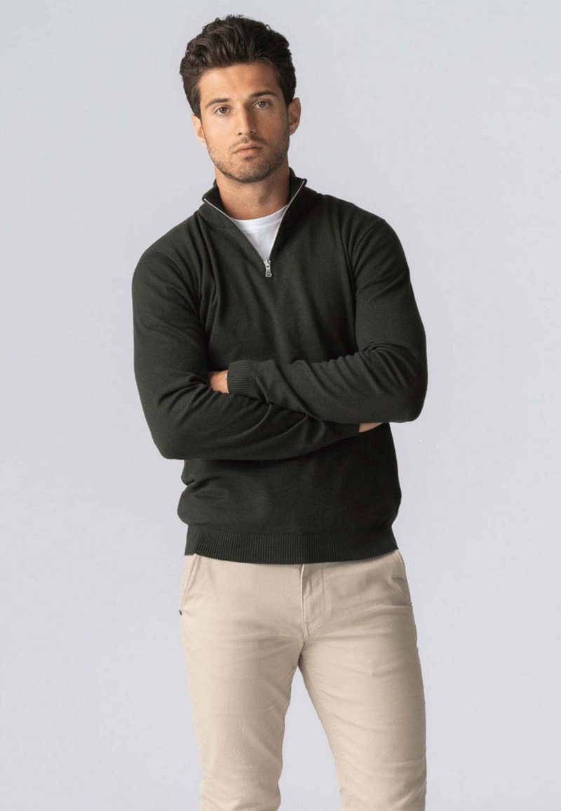 Mørkegrøn sweater med lynlås ved halsen, lavet af strikstof med ribbede manchetter og kant, kombineret med lysebeige bukser, sat mod en ensfarvet baggrund.