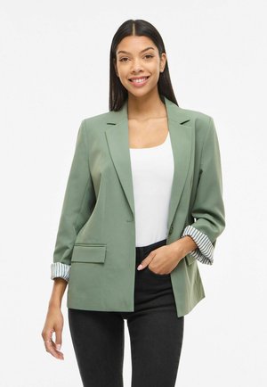 Glunderende vrouw met lang donker haar draagt een lichtgroene blazer over een wit shirt en zwarte broek, met één hand in haar zak, staand tegen een witte achtergrond.
