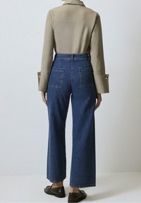 Jeans z wysokim stanem w ciemnoniebieskim kolorze, w szerokiej nogawce, z dwoma tylnymi kieszeniami i teksturowanym wykończeniem z bawełny. Połączone z beżową koszulą z kołnierzykiem.