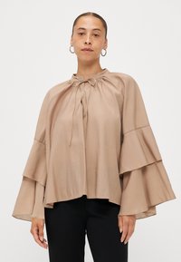 Beige, lightweight blouse with a high neckline, drawstring detail, and bell sleeves featuring layered ruffles. Smooth texture, relaxed fit.