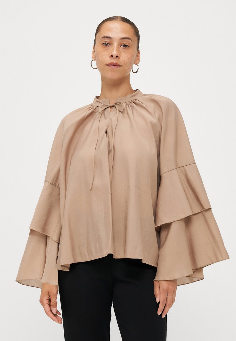 Beige, lightweight blouse with a high neckline, drawstring detail, and bell sleeves featuring layered ruffles. Smooth texture, relaxed fit.