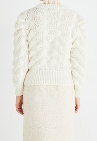 Pull en maille blanche épaisse avec motifs en zigzag, poignets et ourlet côtelés, associé à une jupe crème douce et texturée.