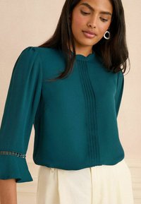 Tyrkysová blúzka s plisovaným dizajnom, rozšírenými rukávmi a ozdobným lemovaním na manžetách. Hladká textúra s skromným výstrihom.