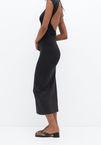 Robe noire sans manches avec un dos nu, silhouette ajustée et ourlet longueur cheville. Texture lisse, design minimaliste, portée avec des sandales à glissière noires.