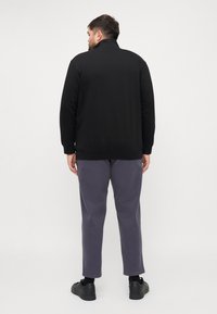 Sudadera negra con cuello alto y dobladillo acanalado, combinada con pantalones grises holgados. El modelo está de espaldas a la cámara, mostrando un diseño sencillo.