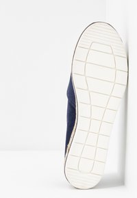 Marinblå mockaskor med en texturerad vit gummiyttersula. Yttersulan har ett rutmönster och är fäst vid en mellansula med en beige accent.