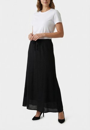 Donna con indosso una camicia bianca a maniche corte e una gonna lunga nera con coulisse, in piedi con le mani giunte davanti, indossa tacchi neri.