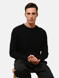 Maglione nero con scollo a costine, realizzato in soffice tessuto testurizzato. Il modello è seduto su uno sgabello di legno, indossando pantaloni neri. Design semplice e aderente.