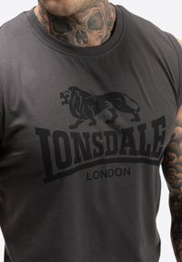 Siva majica bez rukava s velikim crnim grafikom lava i natpisom "LONSDALE LONDON". Izrađena od glatkog materijala s dizajnom okruglog izreza.