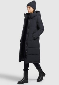 Abrigo acolchado negro con capucha, patrón de acolchado, cremallera frontal y bolsillos laterales, combinado con botas negras y un gorro de punto.