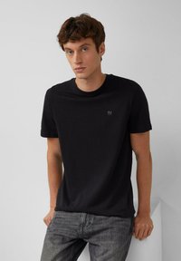 Zwarte katoenen T-shirt met korte mouwen en een ronde hals. Subtiel logo-detail op de borst. Gecombineerd met grijze jeans, casual gedragen.