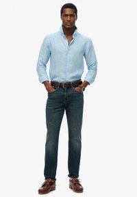  Lichtblauw linnen overhemd met een kraag, gecombineerd met donkere denim jeans. Een bruine leren riem en bordeauxrode veterschoenen maken de look compleet.