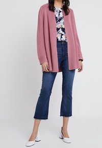 Cardigan texturizzato rosa chiaro sopra una blusa floreale, abbinato a jeans blu tagliati e scarpe con tacco bianco.