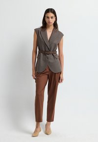 Kvinde iført en ærmeløs brun blazer med bælte, brune tapered bukser, beige hæle, stående mod en ensfarvet hvid baggrund.