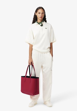 Polo in misto cotone color avorio, abbinata a pantaloni coordinati. Borsa tote rossa con rifiniture nere. Sciarpa verde con motivo legata al collo.