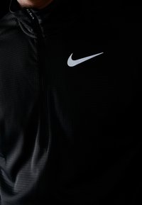 Musta vetoketjullinen takki, joka on valmistettu teksturoidusta kankaasta. Vasemmassa rintakehän kohdassa on harmaa Nike-logo. Kangas vaikuttaa kevyeltä ja sileältä.