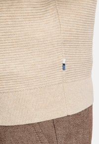 Beige Strickpullover mit geripptem Muster, ausgestattet mit einem einfarbigen Saum und einem kleinen Akzent-Tag mit blauen und weißen Streifen an der Seitennaht.