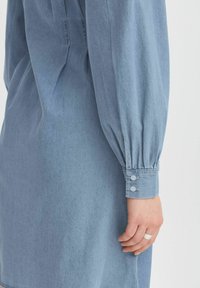 Denim haljina u svijetloplavoj boji s opuštenim krojem, dugim napuhanim rukavima, dugmadi na manžetama i glatkom teksturom.