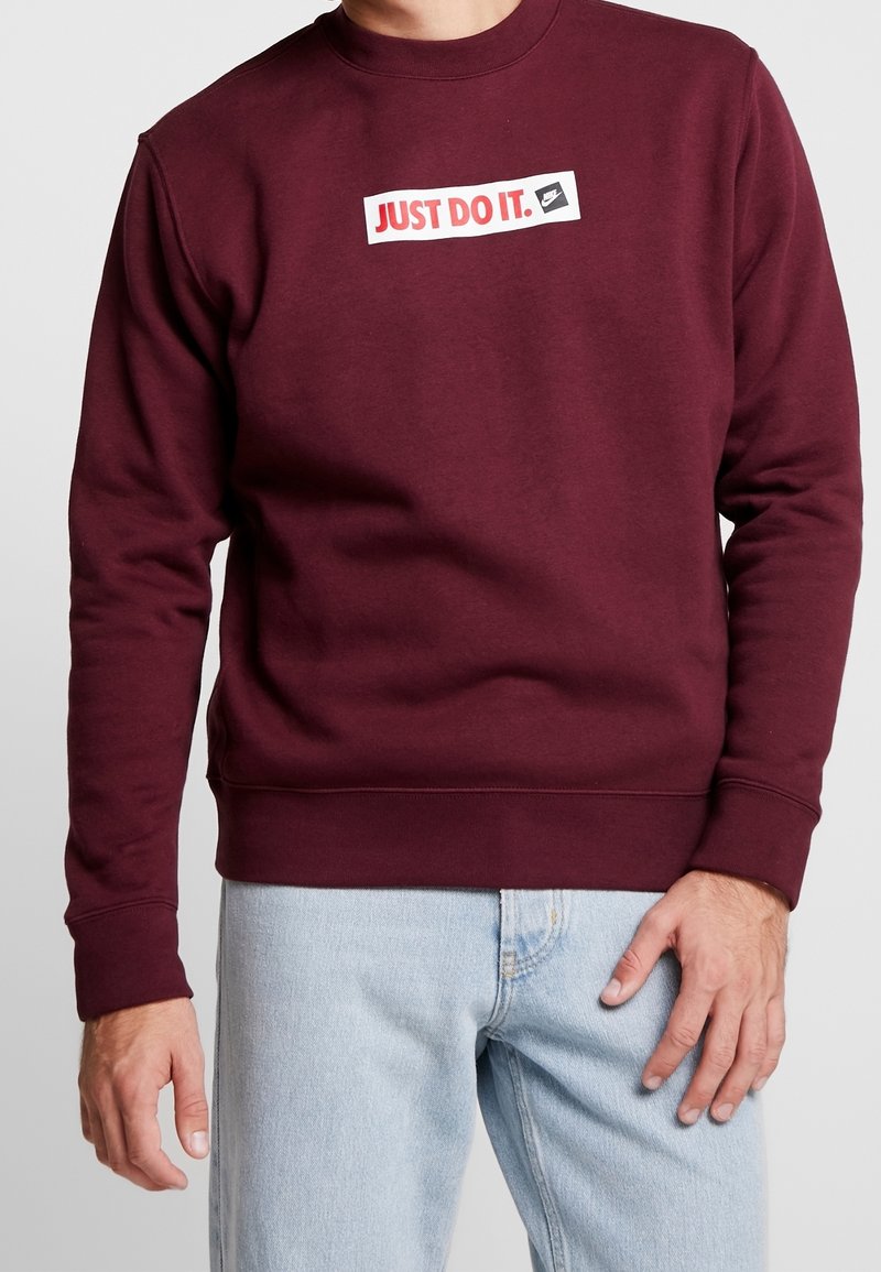 Burgundy sweatshirt i mjukt tyg, med en vit rektangulär lapp med texten "JUST DO IT." i rött och en logotyp, ribbade muddar och nederkant.